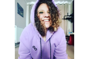 Jodie is smiling at the camera. Brown curly hair with blonde tips spills about her face and she is wearing sparkly gold eyeshadow. She wears a purple Essential hoodie with the hood up.
