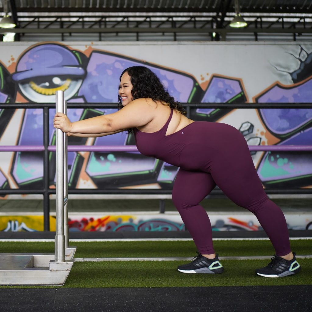 Tarnn is pushing a sled in a gym wearing our Essential Jumpsuit in Burgundy